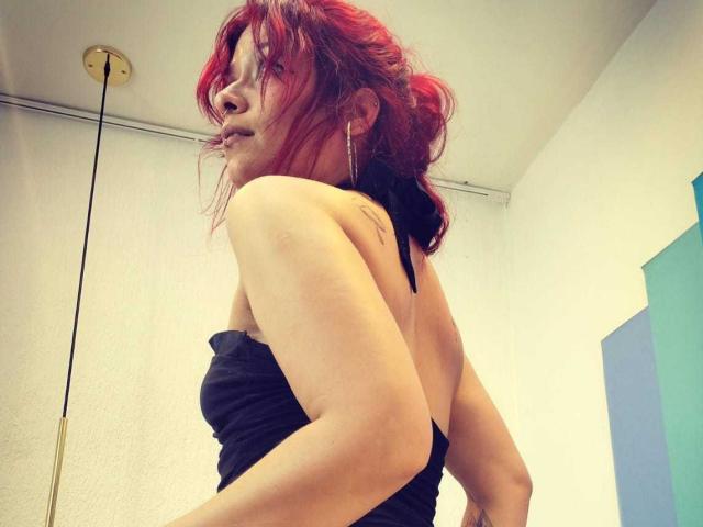 LolyYunno, vista lateral, cabello rojo, top negro sin tirantes, hombros descubiertos, piel clara, tatuaje discreto en el brazo, ligera elevación. mirada, luz natural, ligera sombra, ambiente interior, informal, pose ligeramente provocativa..