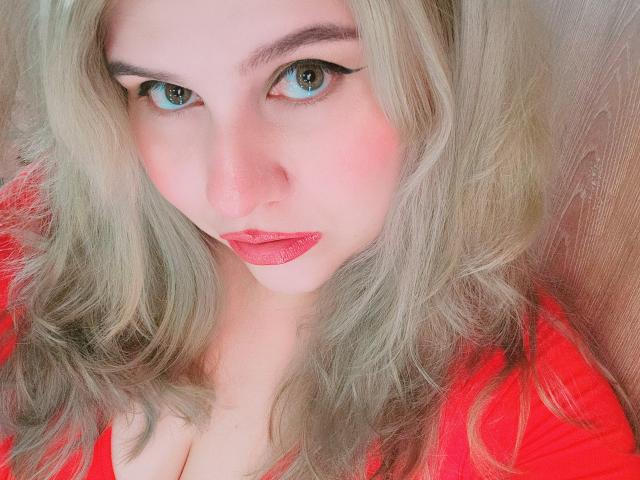 Nahaufnahme von Akkanee mit hellblondem, gewelltem Haar, blauen Augen und rotem Lippenstift. Akkanees intensiver Blick, die leicht geöffneten roten Lippen und die weiche Textur erzeugen eine sexuelle Spannung. Heller roter Hintergrund.