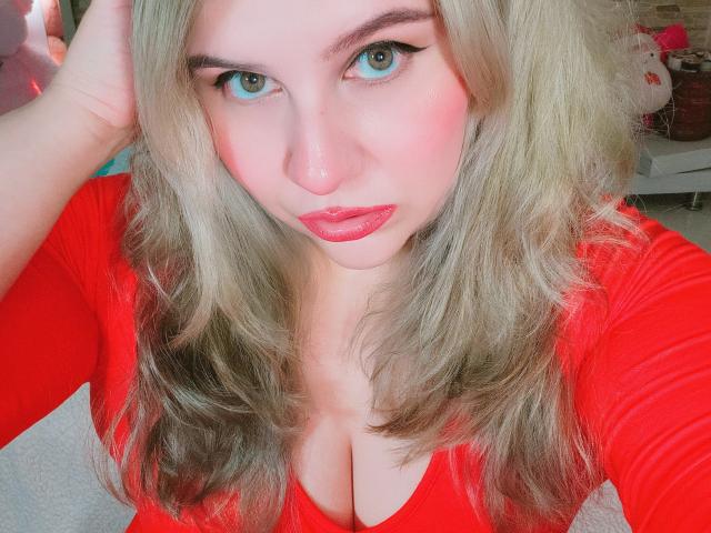 Nahaufnahme von Akkanee mit heller Haut und blauen Augen, die ein rotes Oberteil mit tiefem Ausschnitt, blondes, welliges Haar und rote lippenstift. Akkanee blickt direkt in die Kamera und hat einen verführerischen Ausdruck..