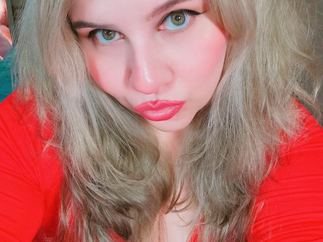 Close-up of Akkanee with blonde, wavy hair and blue eyes, wearing a bright red sweater, pouting with pink lips. Bright colors, intense gaze, and slight sexual tension. Vivid, provocative portrait.