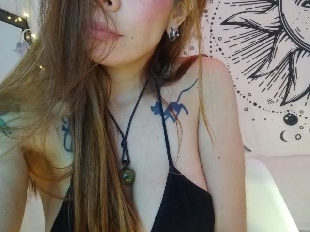 Primer plano del rostro y la parte superior del pecho de AriiannaHot, mostrando cómo se muerde el labio y una peca justo encima. Su cabello castaño cubre parte de su rostro, y tiene un tatuaje negro en el hombro. Increíblemente atractivo/a..