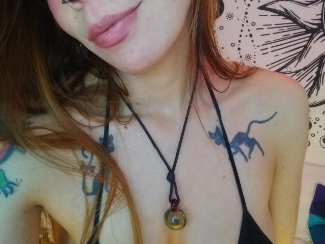 Close-up photo of AriiannaHot, focusing on her face and upper chest. She has full lips, a nose piercing, and tattoos of a dinosaur and flowers. Wearing a black halter top, necklace with a green pendant. Sexual tension evident.