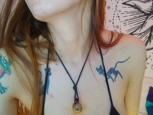 Primo piano di AriiannaHot con i capelli castani, che indossa una collana nera con un ciondolo circolare. Tatuaggi visibili: un drago verde sulla spalla sinistra e un ragno nero sulla spalla destra. Tensione sessuale.