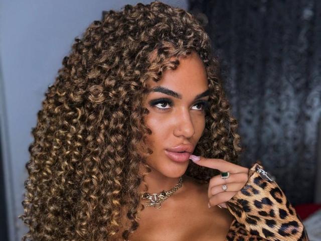 Fotografia de ExoticTala com cabelos castanhos grossos e encaracolados, vestindo uma blusa com estampa de leopardo, com o dedo nos lábios, olhar sedutor, corpo completo. lábios, pele bronzeada, colar de ouro, anéis, pose sexy e confiante..