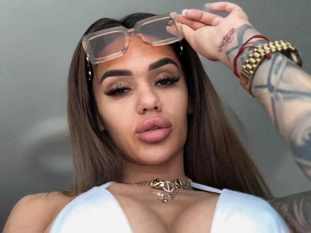 Photograph of ExoticTala, with long brown hair, full lips, and tattoos on her arm. She wears a white top, gold necklace, and glasses on her head, looking at the camera. Her wrist has a gold bracelet.