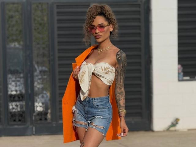 ExoticTala, con cabello rizado, usando gafas de sol rojas, una camiseta blanca anudada, pantalones cortos vaqueros rotos y una chaqueta naranja. por encima del hombro. Brazo tatuado, pose segura, tensión sexual, ambiente urbano..