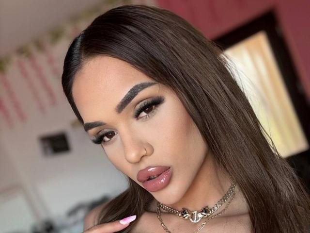 Close-up van ExoticTala, een aantrekkelijke vrouw met lang, steil bruin haar, donkere, rokerige oogmake-up, glanzende roze lippen, en een zilveren ketting met een hartbedel. Ze kijkt verleidelijk naar de camera..