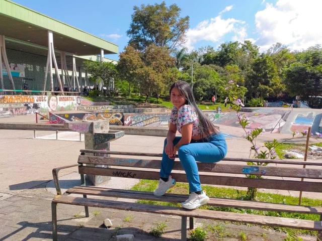 SofiiaThooner está sentada em um banco de madeira em um parque ensolarado, vestindo jeans azuis e uma camisa branca, com um rabo de cavalo comprido. Grafites coloridos ao fundo. Sombras no rosto dela..