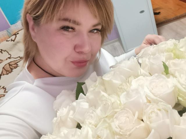 CurlyDreams, avec ses cheveux bruns clairs et sa peau claire, tient un grand bouquet de roses blanches près de son visage. Elle porte une chemise blanche, avec un sourire en coin, et une tension sexuelle palpable..