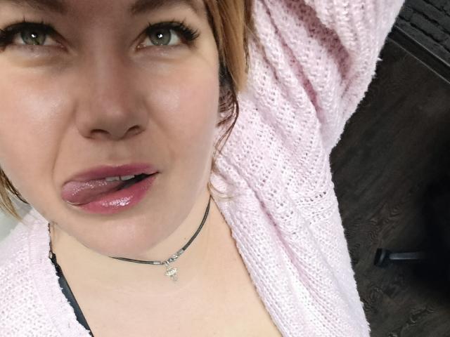 Gros plan sur CurlyDreams, une jeune femme à la peau claire et aux cheveux roux, qui tire légèrement sa langue. Elle porte une serviette rose par-dessus un vêtement noir, et un collier en argent. Ses yeux sont à moitié ouverts, ses lèvres sont brillantes..