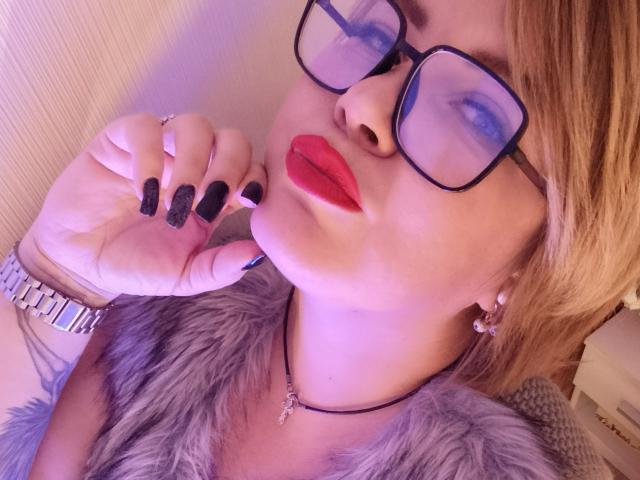 Photo en gros plan de CurlyDreams, avec du rouge à lèvres rouge, du vernis à ongles noir et des lunettes, mordant son doigt. Ils portent un col en fourrure grise, une montre argentée et des boucles d'oreilles. Un look incroyablement sexy..