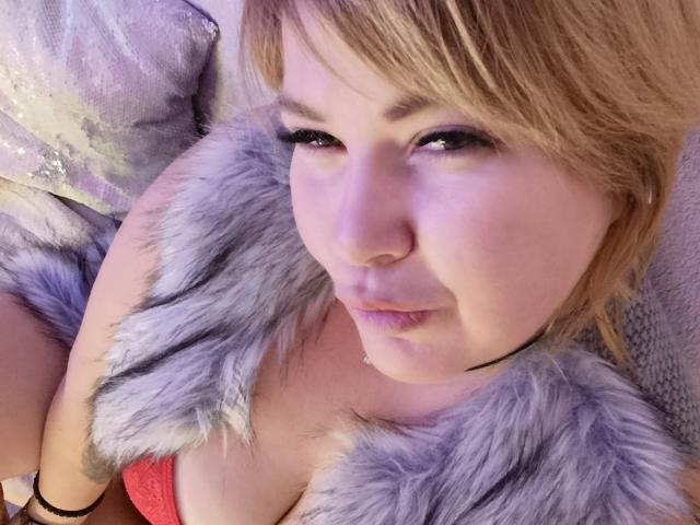 Gros plan de CurlyDreams, avec des cheveux blonds clairs, portant un soutien-gorge rouge et un col en fourrure gris, regardant directement l'appareil photo. Avec les lèvres légèrement entrouvertes, dégageant une tension sexuelle. Éclairage doux.