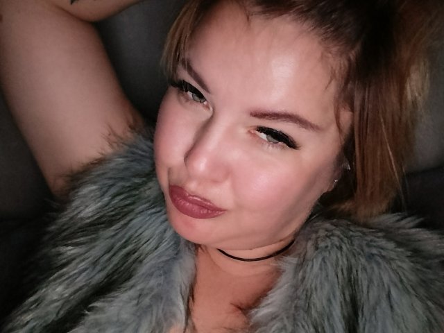 Fotografia ravvicinata di CurlyDreams, una donna bianca con capelli castano chiaro, che indossa un cappotto di pelliccia grigia, con le braccia alzate sopra la testa. Guance leggermente arrossate, un sorriso malizioso con rossetto rosso scuro, sguardo intenso..