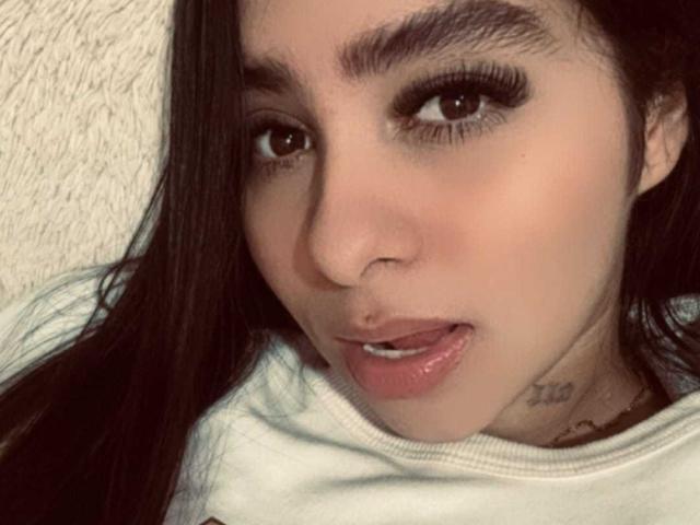 Primo piano di ValeryyRose, una giovane donna con lunghi capelli neri, sopracciglia folte e grandi occhi scuri. Sta portando fuori la lingua, mostrando un piccolo tatuaggio sul collo. Indossa una camicia bianca. La sua espressione è sensuale e provocante..
