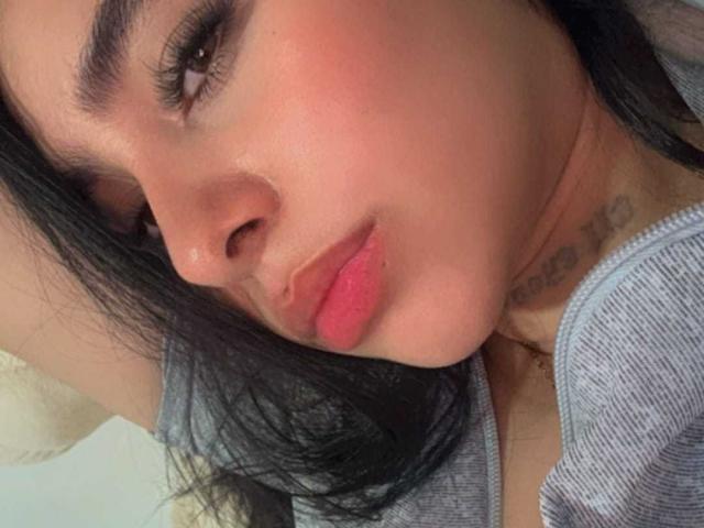 Gros plan de ValeryyRose, avec une peau de couleur moyenne, des cheveux noirs et des lèvres rouges. Le visage de ValeryyRose est légèrement incliné vers la gauche, révélant un tatouage sur le cou. Elle porte un haut gris. Incroyablement sexy..