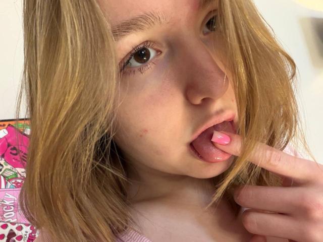 Foto de primer plano de Rima Pecinovsky, rubia, de piel clara, lamiendo el esmalte rosa de sus uñas con un dedo, mirada seductora, cabello despeinado. cubriendo parcialmente el rostro, fondo vibrante de color rosa y blanco, sugerente, tensión sexual, provocativo..