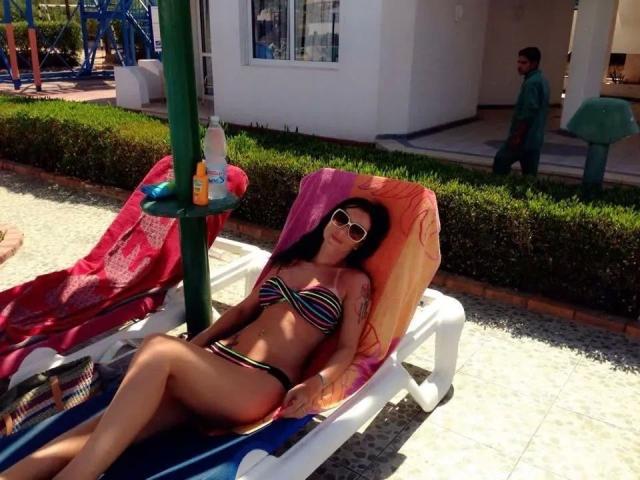 NaomiStarX ligt ontspannen op een ligbed, gekleed in een zwart-wit gestreepte bikini, met een zonnebril en een piercing in haar navel. Zongebruind met gespreide benen, waardoor er zichtbare seksuele spanning ontstaat. Helder zonlicht, rode handdoek.