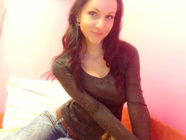 NaomiStarX - attrayante, cheveux bruns et longs, ondulés, haut noir à manches longues, léger sourire, penchée en avant, main sur la cuisse, jean. jeans, lumière douce, fond rose et blanc, tension sexuelle subtile, pose décontractée, attrayant.