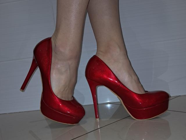 Primo piano dei piedi di Amberia, che indossa scarpe rosse lucide con tacchi a spillo. Le sue gambe sono leggermente divaricate, il che mette in risalto gli archi plantari e la superficie riflettente sotto i suoi piedi. Di colore rosso acceso, con una texture lucida..