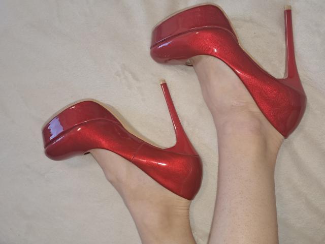Las piernas de Amberia, pálidas y lisas, lucen unos tacones rojos brillantes y altos, con un diseño de aguja, y los pies están ligeramente separados, de color rojo. los tacones reflejando la luz, creando una atmósfera provocativa y sensual..