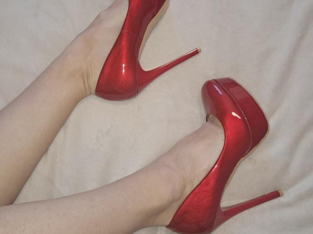 Nahaufnahme von Amberias Beinen, die glänzende, rote Stiletto-High Heels tragen, die Füße sind leicht gekreuzt, blasse Haut, rot. Die Absätze glänzen, die Textur der Absätze ist glänzend, eine sexy Ausstrahlung, eine verführerische Pose..