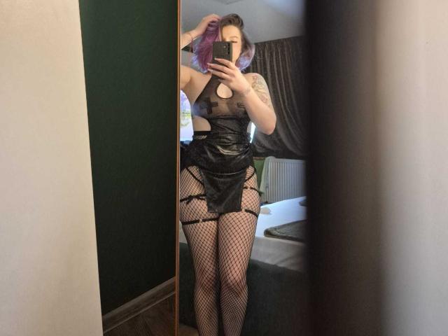 NatashaBlaze, debout devant un miroir, se prend en photo. Elle portait une tenue en latex noire et transparente, avec des découpes, des bas en filet et des tatouages sur son bras droit. Cheveux violets, tension sexuelle
.