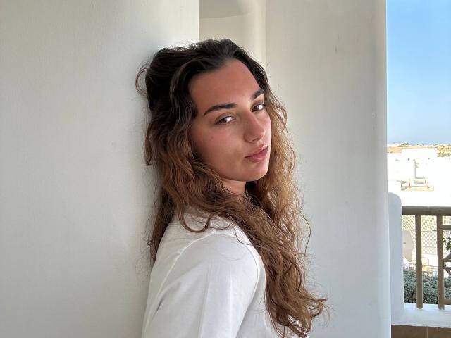 Vivien Henifin, eine kurvige Frau mit langen, welligen braunen Haaren, lehnt sich an eine weiße Wand und trägt ein weißes Oberteil. Sie blickt mit einem verspielten, verführerischen Ausdruck über ihre Schulter. Das Sonnenlicht betont ihr Gesicht..