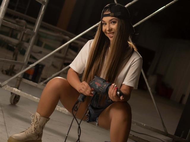 IrisMia est assise sur un échafaudage métallique, les jambes écartées, et tient une perceuse électrique. Portant une casquette à l'envers, un t-shirt blanc, un short en jean et des bottes à lacets. Cheveux tressés, regard intense, séduisant.