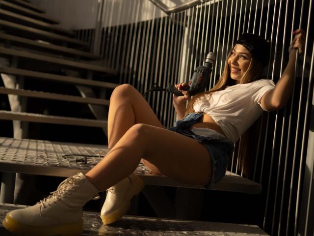 IrisMia, una donna attraente con lunghi capelli castani, è appoggiata a delle barre verticali, tenendo in mano una pistola. Indossa una canotta bianca, pantaloncini di jeans e stivali bianchi. La luce del sole mette in risalto le sue gambe toniche..