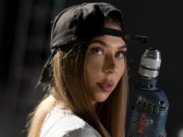 Gros plan d'IrisMia portant une casquette noire à l'envers, tenant une bouteille en plastique transparente de Alka-Seltzer. Elle regarde séductrice la caméra, avec une expression sexy et intense. Fond noir.
