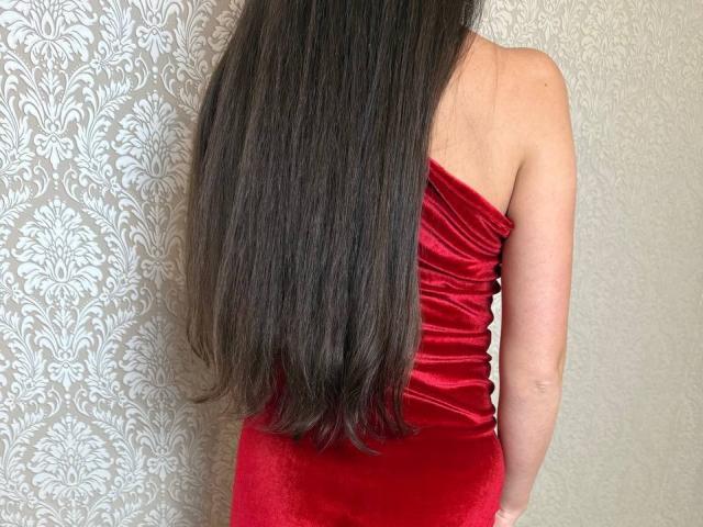 Rouzi est de retour, avec ses longs cheveux noirs, vêtue d'une robe rouge en velours moulante sans bretelles, une légère courbure dans le dos, une peau claire. Mur blanc orné de motifs floraux sur la gauche, ambiance sexy et séduisante..