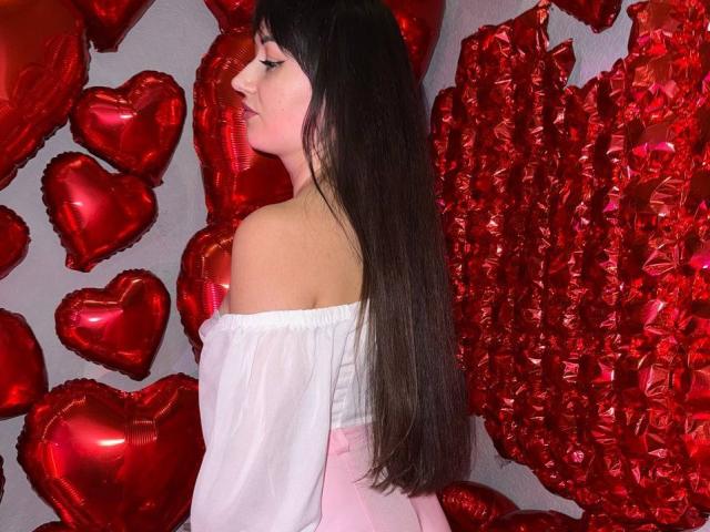 Rouzi, avec de longs cheveux noirs, tournée vers la gauche, portant un haut blanc à une épaule, entourée de ballons en forme de cœur rouges, couleur rouge intense, tension sexuelle, contraste entre le haut blanc et les cœurs rouges..