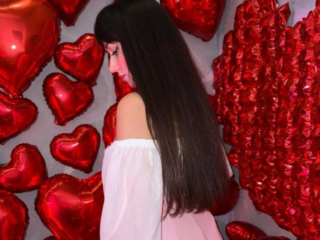 Rouzi, avec de longs cheveux noirs, un haut blanc à une épaule, tournée vers la gauche, entourée de ballons rouges brillants en forme de cœur. La tension sexuelle dégagée par le profil de Rouzi et les cœurs rouges vifs..