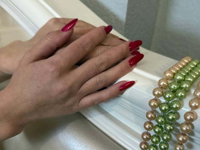 Rouzi's hands with bright red, long, pointed nails rest on a white surface. Beaded necklace with green and gold pearls lies nearby, reflecting light. Tense, sensual atmosphere.