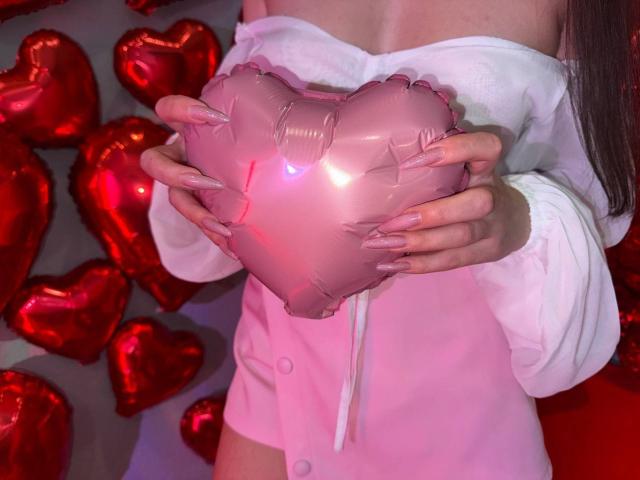 Rouzi tiene tra le mani un palloncino a forma di cuore rosa lucido, indossando una camicia bianca. Oggetti a forma di cuore rossi sono sparsi sullo sfondo. Texture luminosa e lucida sui cuori. Tensione sessuale.