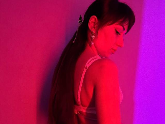Rouzi with dark hair in a ponytail, wearing a white bra, illuminated by intense red and purple lighting. She's turned to the side, eyes closed, creating a sultry, sexy vibe.
