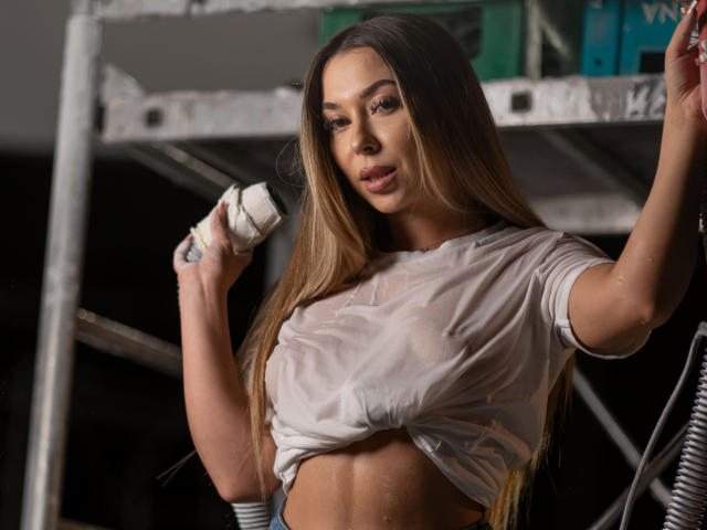 IrisMia, une femme séduisante avec de longs cheveux bruns, tient un microphone, vêtue d'un crop top blanc mouillé qui épouse ses formes. des seins et des abdominaux bien dessinés. Son expression est sensuelle et confiante..