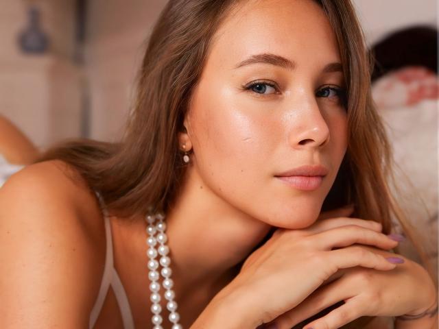 Close-up of AmmaLoyXX, brown-haired woman with light skin, blue eyes, and subtle makeup, wearing a pearl necklace, leaning on her hands with a seductive expression, soft lighting, blurry background.