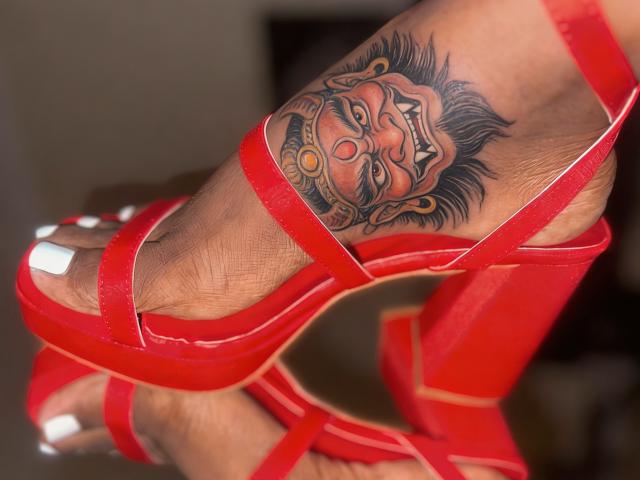 Close-up van een been met een bruine huid, met een kleurrijke tatoeage van een lachende Boeddha op de binnenkant van de dij. CcamillaX draagt rode, smalle hoge hakken. De tatoeages zijn opvallend en levendig..