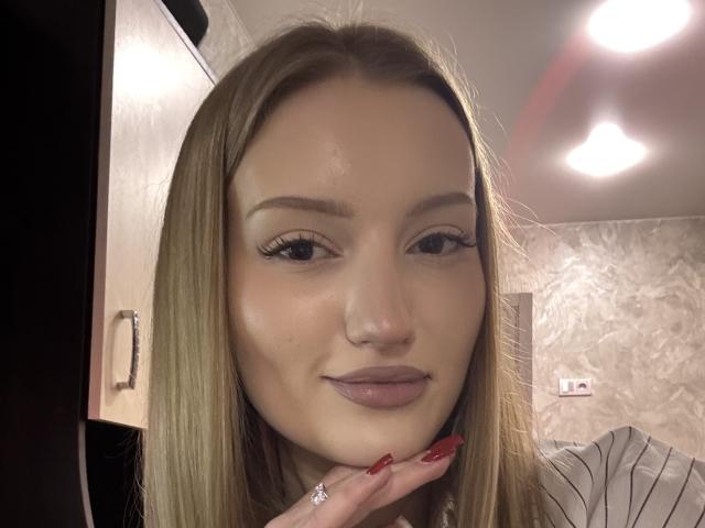 Primer plano de HeilaGr, una mujer de piel clara con cabello rubio largo, labios carnosos y uñas rojas, que apoya su barbilla en su mano. mirando directamente a la cámara, con luces brillantes sobre la cabeza.