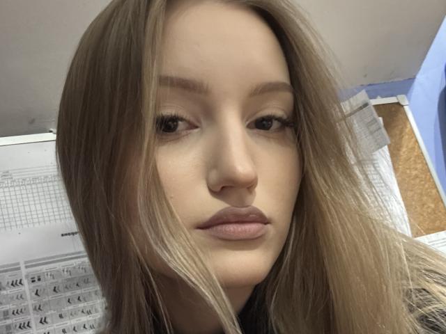 Nahaufnahme von HeilaGr, einer jungen Frau mit hellbraunen Haaren, neutralem Gesichtsausdruck und vollen Lippen. SheilaGr's Gesicht ist leicht zur Seite geneigt, im Hintergrund sind ein Dokument und eine hellblaue Wand zu sehen. Sie blickt direkt in die Kamera mit einem subtilen, fast gleichgültigen Blick..