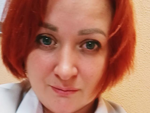 Nahaufnahme eines Fotos von AgilaMipo, einer rothaarigen Frau mit heller Haut und blauen Augen, die direkt in die Kamera schaut. Ein leichtes Lächeln. Ihre Lippen sind leicht geöffnet, was auf sexuelle Spannung hindeutet..