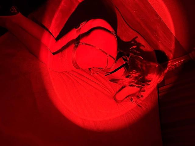 Close-up, high-contrast photo of MiraGi in a red-lit, abstract setting. MiraGi's body is twisted, limbs tangled, with intense red light highlighting curves and shadows. Erotic, intense atmosphere.