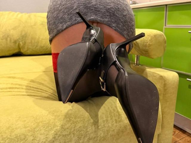 MiraGi está sentada em um sofá amarelo, com os joelhos levantados, vestindo uma saia cinza, saltos altos pretos com tiras nos tornozelos, e com o bumbum à mostra. Roupa íntima vermelha. Armários verde-claros ao fundo. Incrivelmente atraentes..