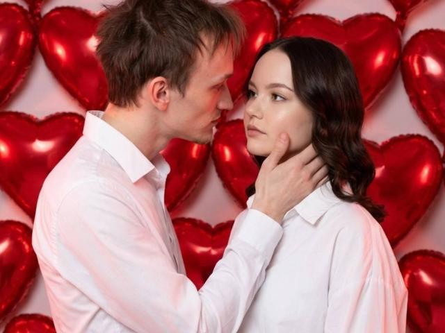 WeksaAndOliver, a guy with short brown hair and WeksaAndOliver, a woman with long black hair, both in white shirts, face each other intimately. Red heart balloons fill the background. His hand grips her neck.