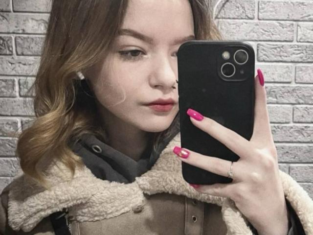 Close-up photo of WeksaAndOliver, fair-skinned woman with wavy brown hair, red lipstick, and red nails, holding a black phone with dual cameras. Wearing a brown coat with a fur collar. Background: textured gray brick wall. Fucking sexy.
