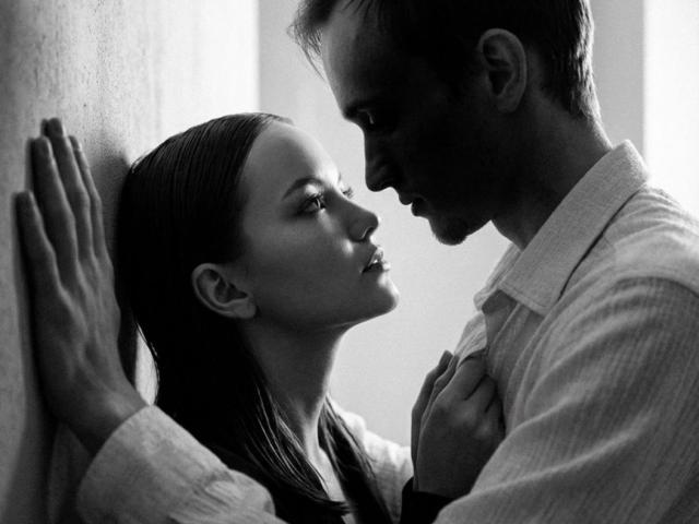 Photographie en noir et blanc de WeksaAndOliver, une femme aux longs cheveux pressée contre un mur, les mains levées, regardant intensément. d'un homme barbu portant une chemise avec un col, le nez presque en contact. Tension sexuelle.