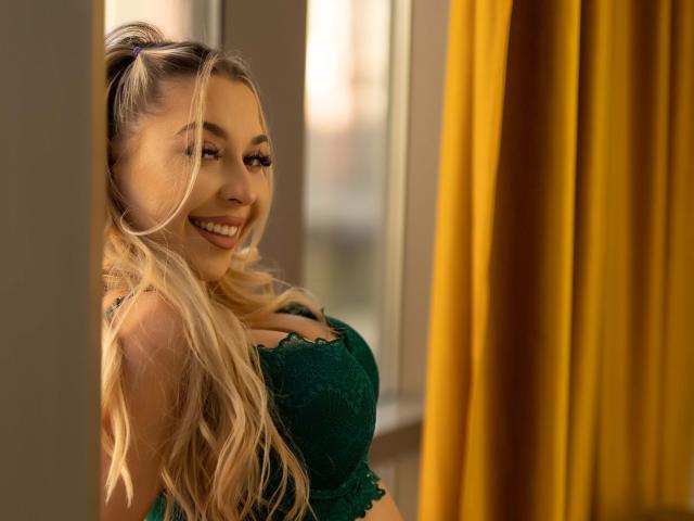 IrisMia with long, wavy blonde hair, smiling seductively, wearing a green lace top that accentuates her large breasts. Yellow curtains in the background. Bright sunlight highlights her face. Fucking hot.