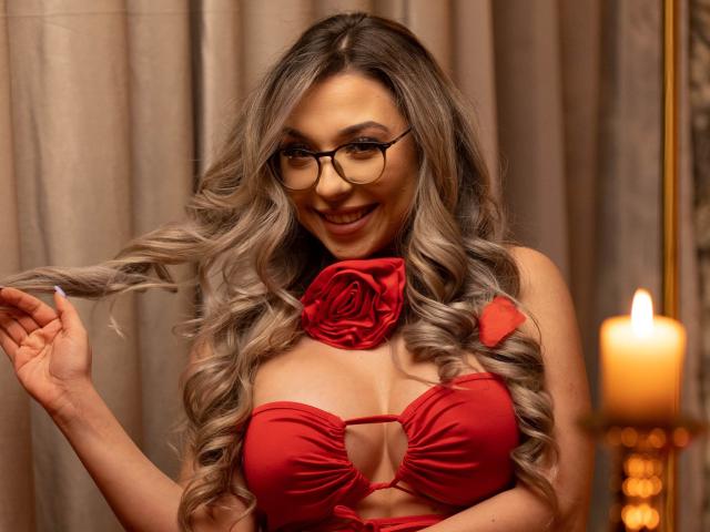 IrisMia, avec de longs cheveux ondulés, gris-blonds, portant un soutien-gorge rouge avec un détail en forme de rose, tenant une mèche de cheveux, souriante. de manière séduisante. L'arrière-plan comprend une bougie allumée sur la droite.