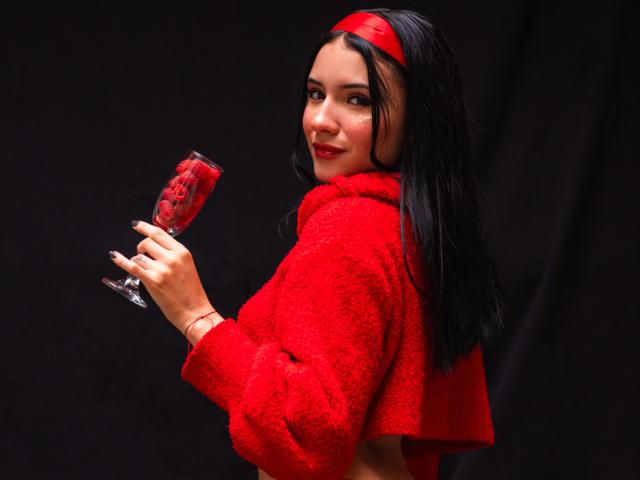 Ilustração digital de LizzeJoy com cabelos longos e pretos, uma faixa de cabeça vermelha e um casaco vermelho felpudo, segurando um copo com um tom avermelhado. Olhando por cima do ombro, fundo preto, pose sexy e confiante..